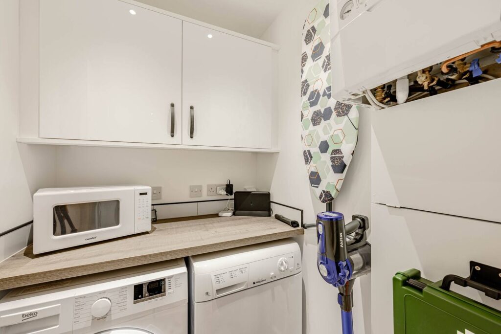 Compact utility room with white cabinets, microwave, washing machine, ironing board, and vacuum cleaner.