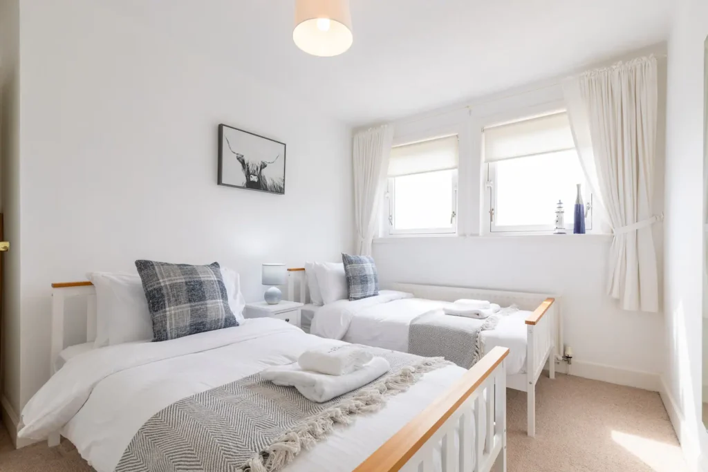 Bright twin bedroom with white decor, cozy bedding, plaid pillows, and large windows letting in natural light.