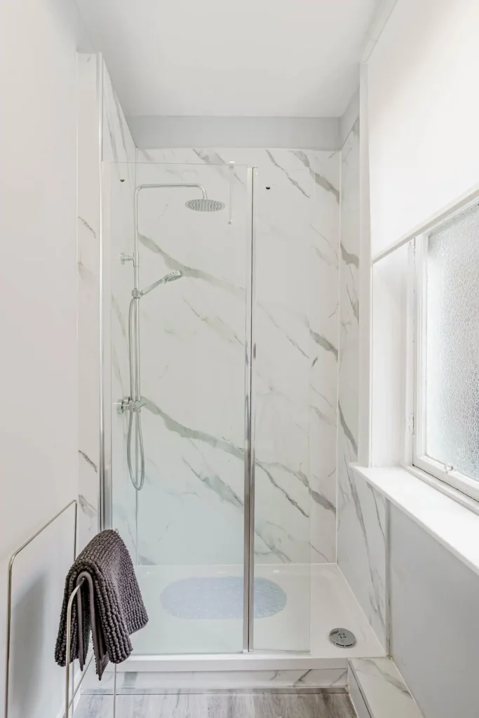 Modern marble shower with glass door and rain showerhead in a bright bathroom. Towel hangs on nearby rack.