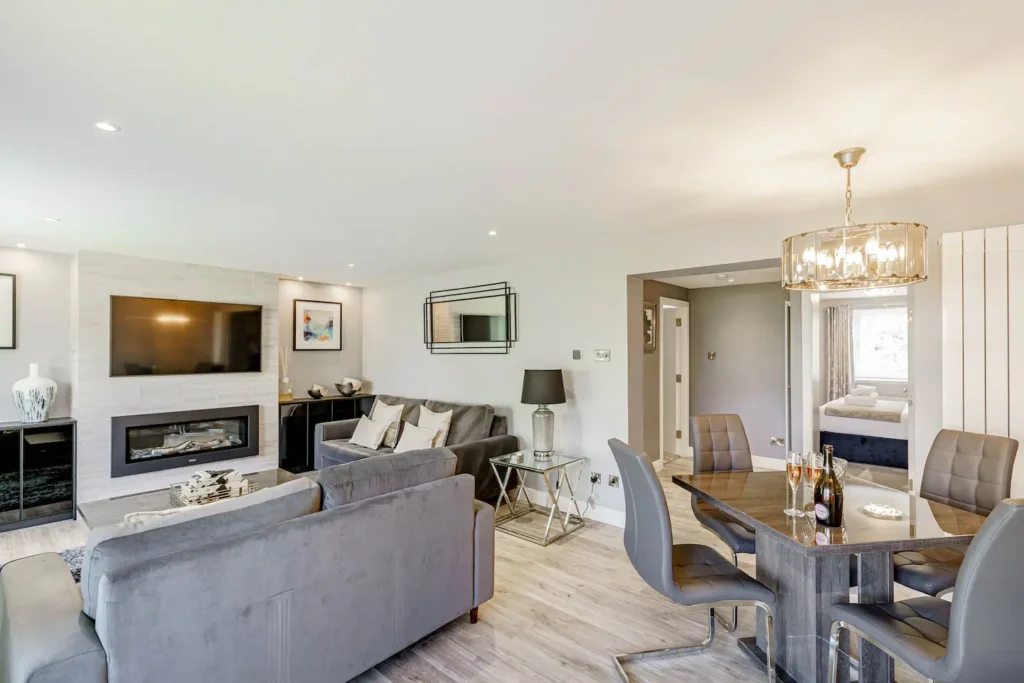 Modern living and dining area with gray sofas, fireplace, wall-mounted TV, and elegant chandelier.