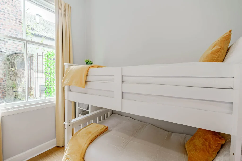 White bunk bed with yellow pillows and blanket in a bright room with large window and beige curtains.