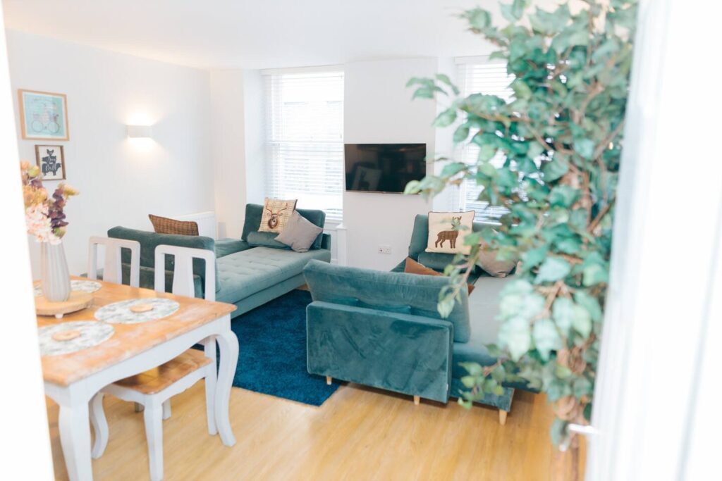 Bright living room with green sofas, dining table, and wall art, featuring a large indoor plant and a mounted TV.