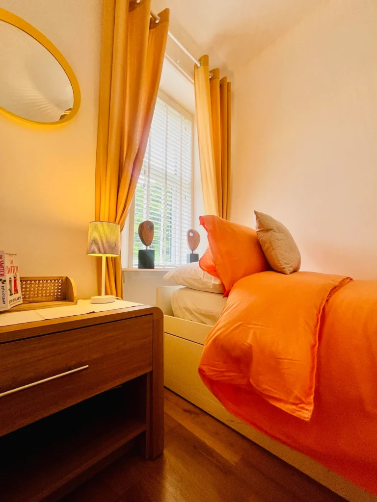 Cozy bedroom with orange bedding, wooden nightstand, and warm lighting by a sunny window with yellow curtains.