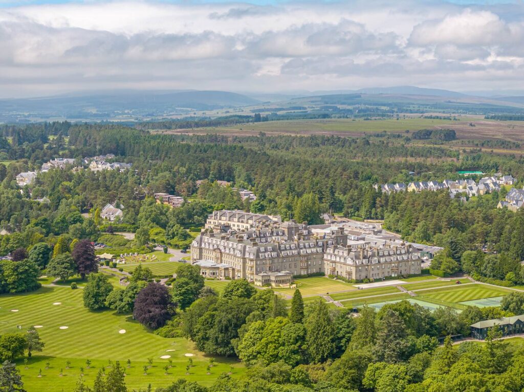 Aerial view of a large historic hotel surrounded by lush greenery and golf course, under a partly cloudy sky.