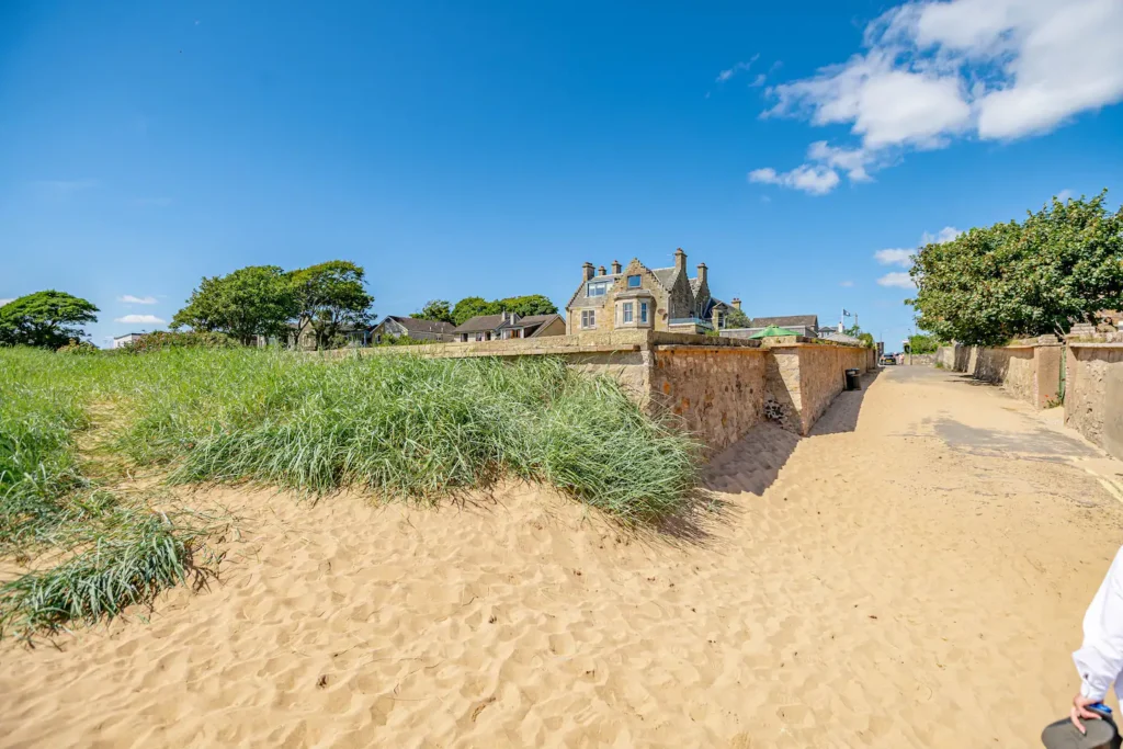 Sandy path leading to a stone house with blue sky and grassy dunes, perfect for a sunny seaside stroll.