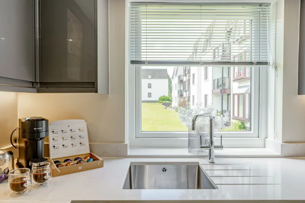 Modern kitchen with a coffee maker, pods, and a sink by a window with blinds overlooking a green lawn and apartments.