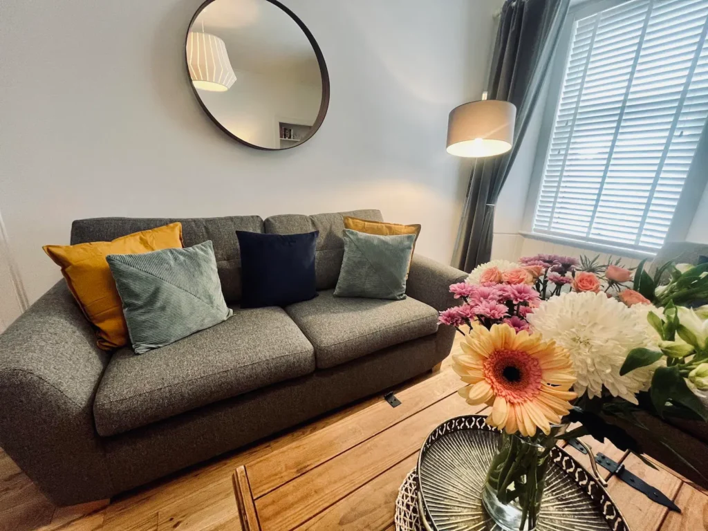 Cozy living room with gray sofa, colorful pillows, flowers on a wooden table, and a round mirror reflecting soft lighting.