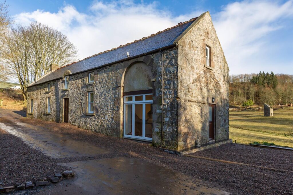 Rustic stone farmhouse with large windows in a serene countryside setting under a blue sky.