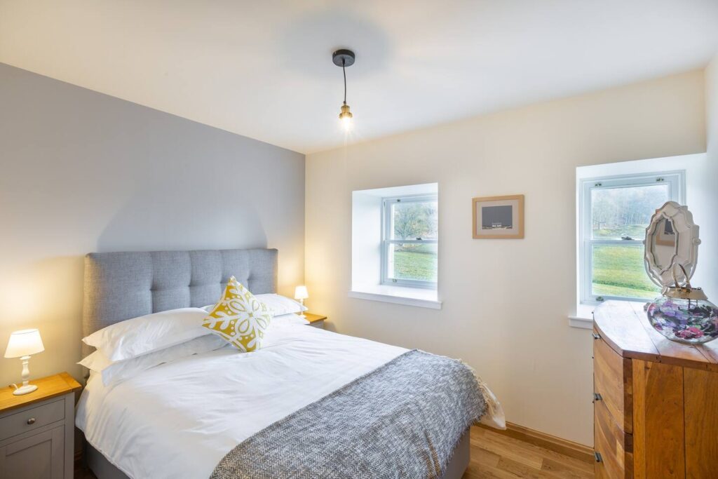 Bright bedroom with double bed, gray headboard, yellow pillow, wooden dresser, and two windows with scenic views.