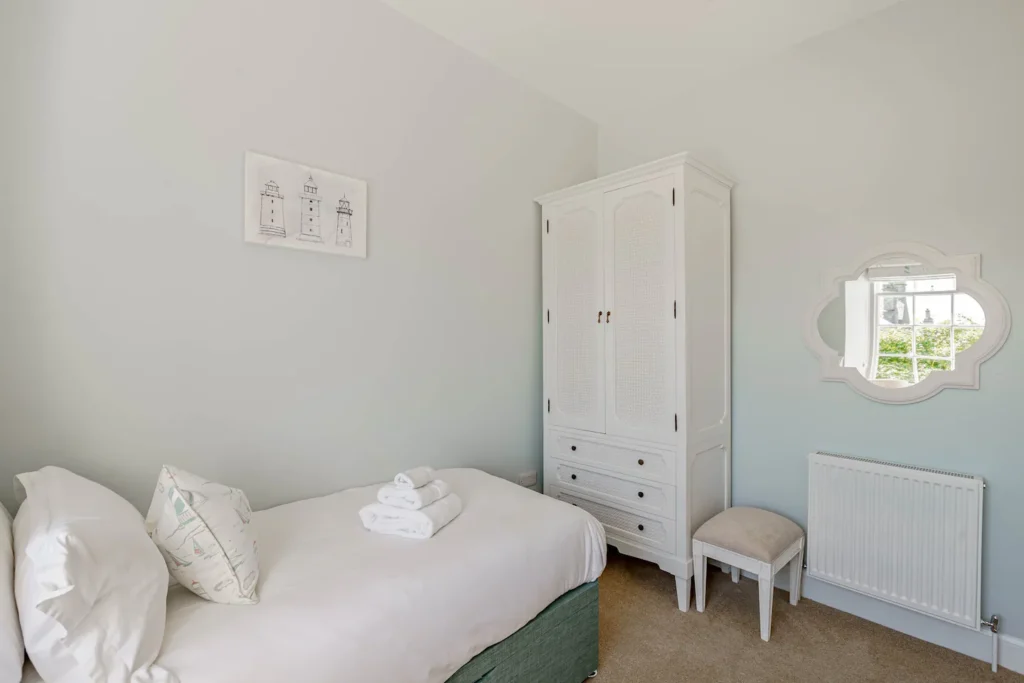 Minimalist bedroom with a single bed, white wardrobe, and mirror, featuring lighthouse artwork on the wall.