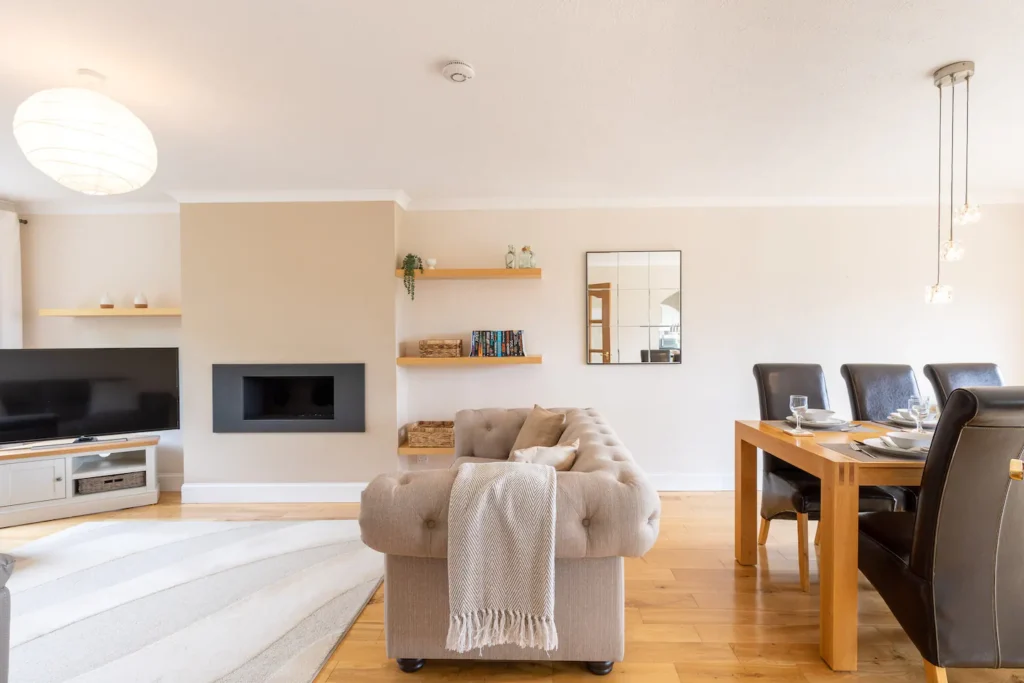 Modern living and dining area with beige sofa, TV, fireplace, and elegant dining set under stylish pendant lights.