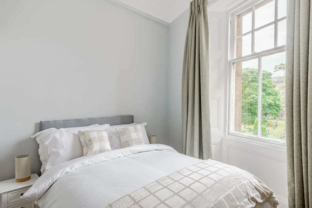 Bright bedroom with cozy bedding, large window, and scenic view. Neutral tones and natural light create a tranquil space.