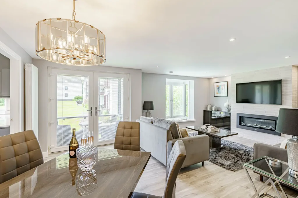 Modern living room with large windows, stylish chandelier, and cozy seating area focused on a sleek fireplace and TV.