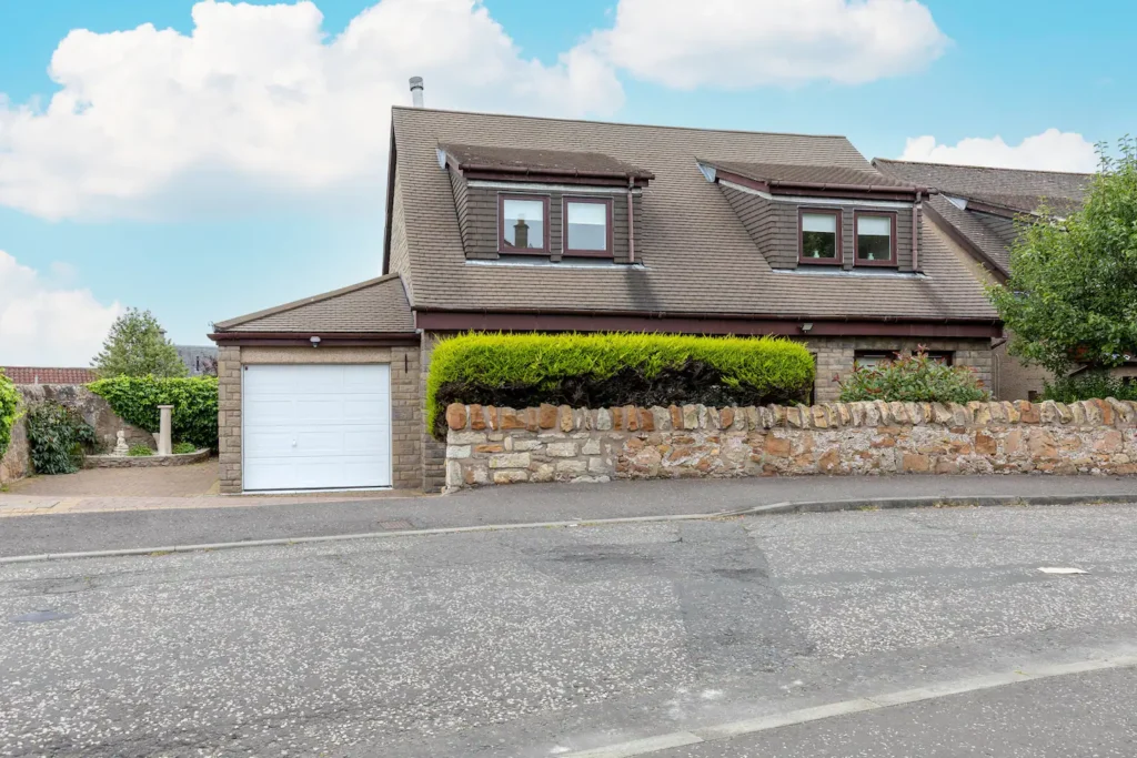 Suburban house with brown roof, garage, and stone wall on a sunny day, surrounded by greenery and blue sky.