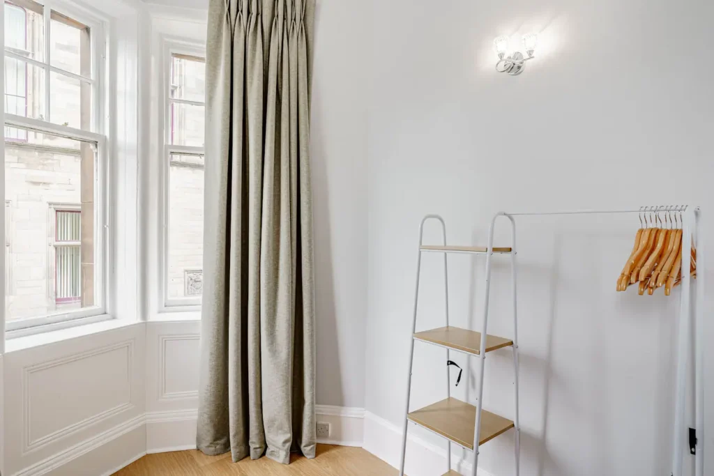 Minimalist room with light curtains and a white ladder shelf with wooden hangers. Bright and modern interior design.