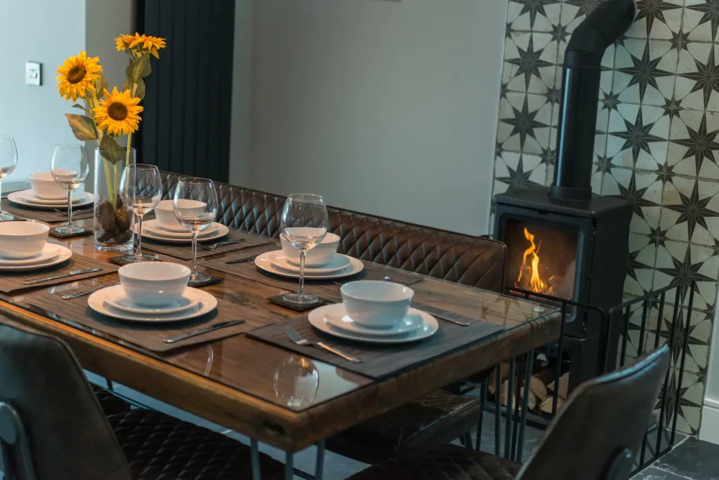 Cozy dining room with wooden table, elegant tableware, sunflowers, and a lit stove by the tiled wall.