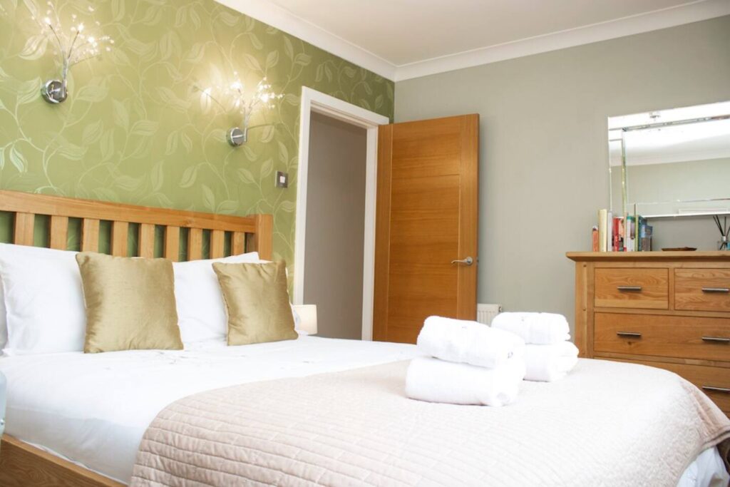 Cozy bedroom with green accent wall, wood bed frame, soft pillows, and neatly folded towels on the bed.