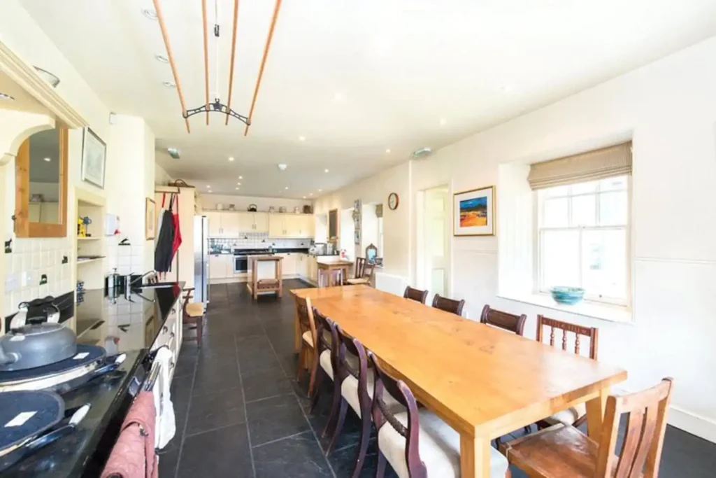 Spacious kitchen with long wooden dining table, modern appliances, and white cabinetry under bright ceiling lights.