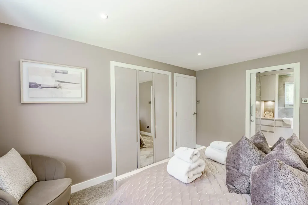 Modern bedroom with mirrored wardrobe, cozy chair, and neatly folded towels on a plush bed.