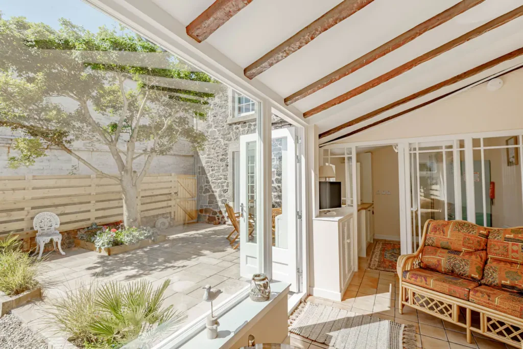 Sunlit conservatory with garden view, open doors, and cozy rattan furniture in a rustic stone-walled home.