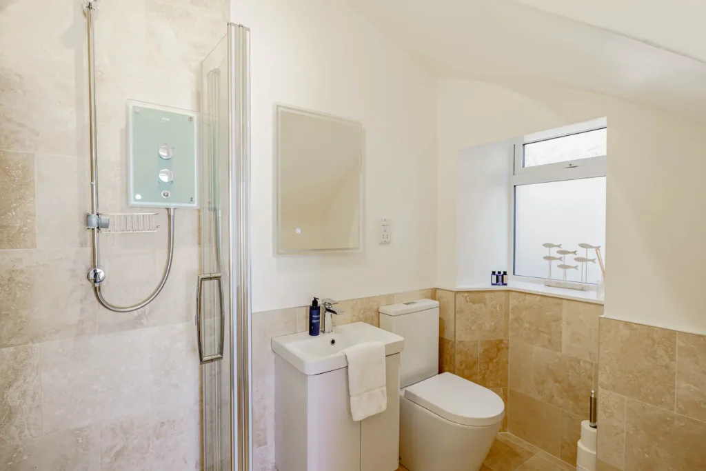 Modern bathroom with glass shower, toilet, and sink. Beige tiles and natural light create a clean, serene atmosphere.