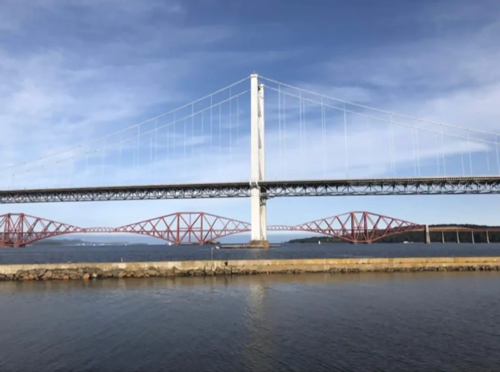 Suspension bridge over calm water with distant red steel bridge under blue sky.