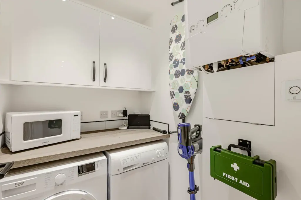 Compact laundry room with washing machine, microwave, ironing board, vacuum, and first aid kit on the wall.