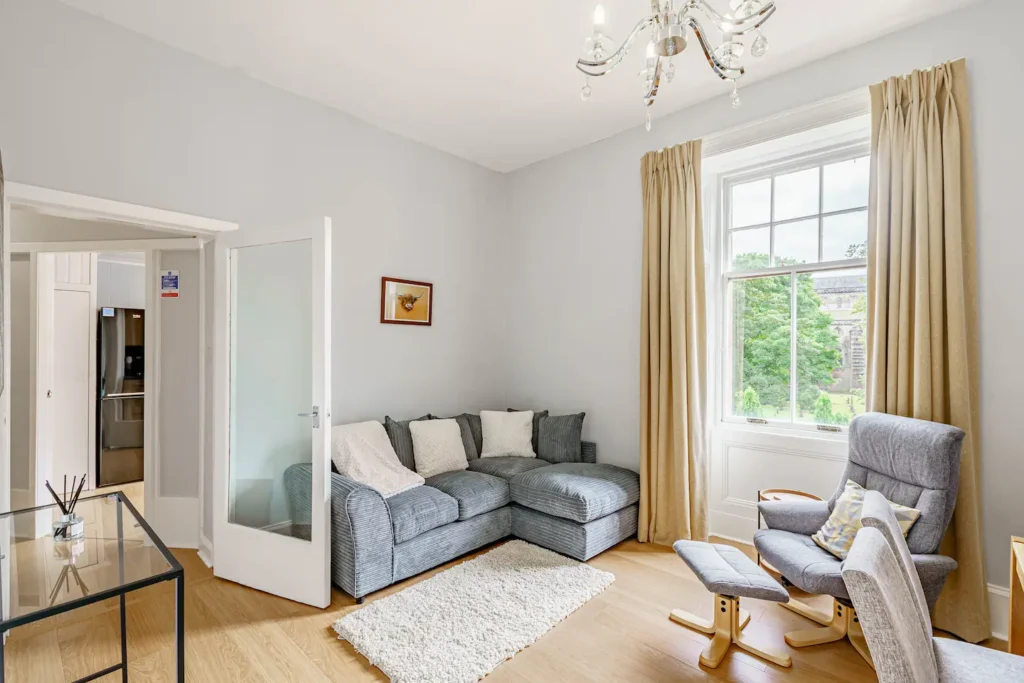 Bright living room with gray sectional sofa, armchair, glass table, and large window with beige curtains.