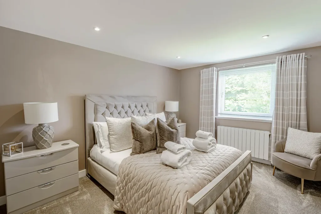 Modern bedroom with elegant decor, featuring a plush bed, bedside lamps, neutral tones, and natural light from a window.