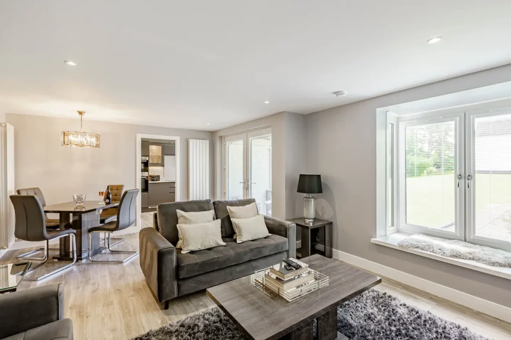 Modern living room with gray sofas, dining area, and cozy decor, featuring large windows for natural light.