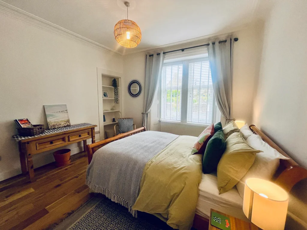 Cozy bedroom with wooden bed, soft lighting, and large window. Decorative table and modern decor complete the inviting space.