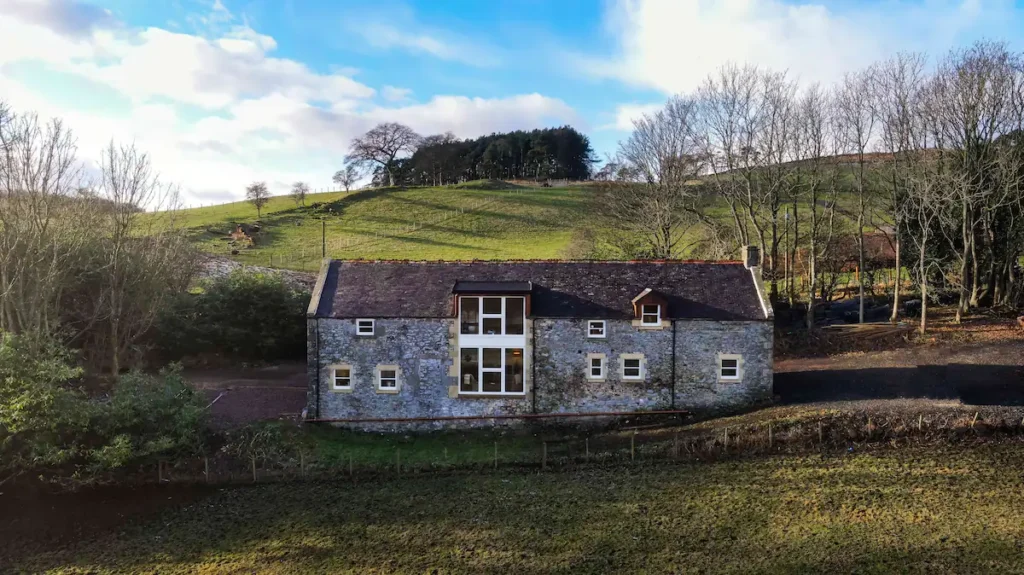 Stone farmhouse nestled in a lush green landscape with blue skies and trees in the background.