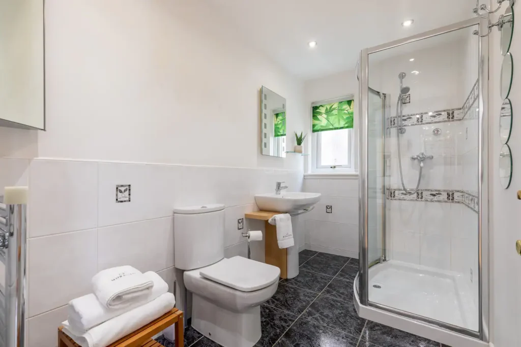 Modern bathroom with glass shower, white tiles, toilet, sink, and green accents.