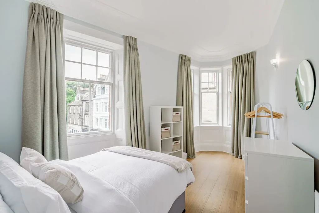 Bright, modern bedroom with large windows, light curtains, and white furniture in a cozy apartment setting.