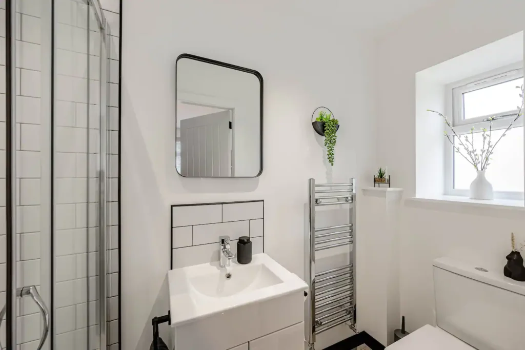 Modern bathroom with white tiles, a square mirror, plants, and a compact sink under natural light.