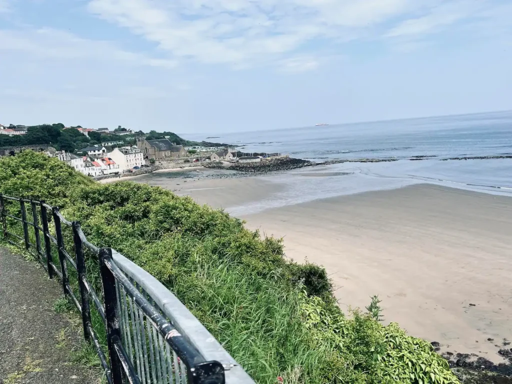 Scenic coastal village view, sandy beach, ocean, lush greenery, and charming hillside houses under a bright sky.