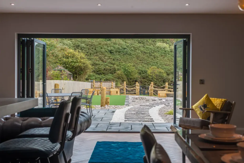 Modern living room with open doors to a scenic garden path, surrounded by lush greenery.