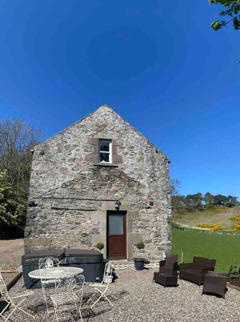 Stone cottage with patio furniture and hot tub, set against a clear blue sky and green landscape.