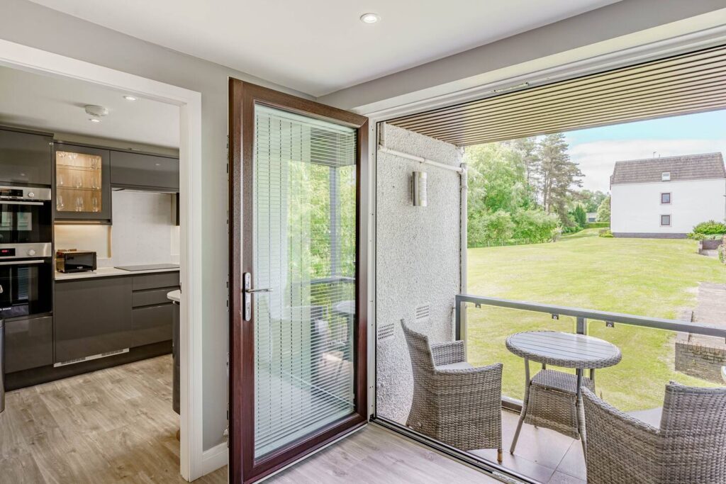 Modern kitchen opening to balcony with wicker furniture, offering garden view through sliding glass doors.