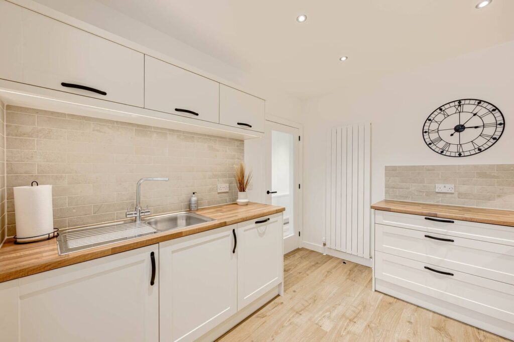 Modern white kitchen with wood countertops, stone backsplash, wall clock, and stainless steel sink.