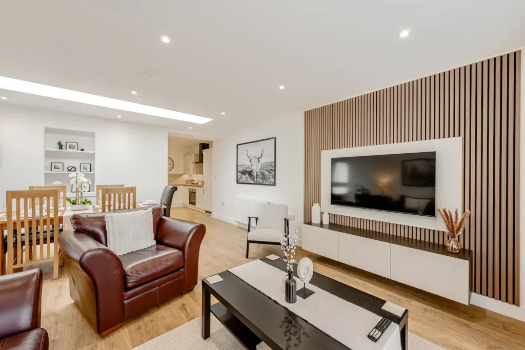 Modern living room with leather chairs, dining area, wall-mounted TV, and open kitchen in a bright, stylish home.