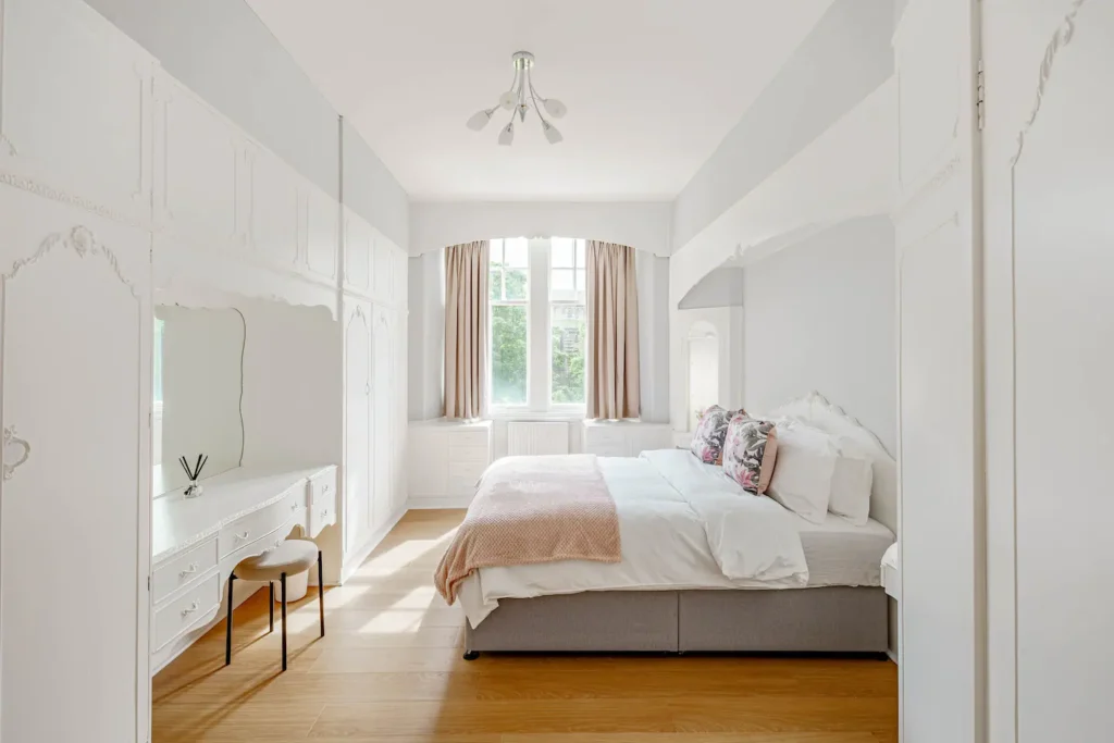 Bright, elegant bedroom with large windows, stylish white furniture, and a cozy bed featuring decorative pillows and a throw.