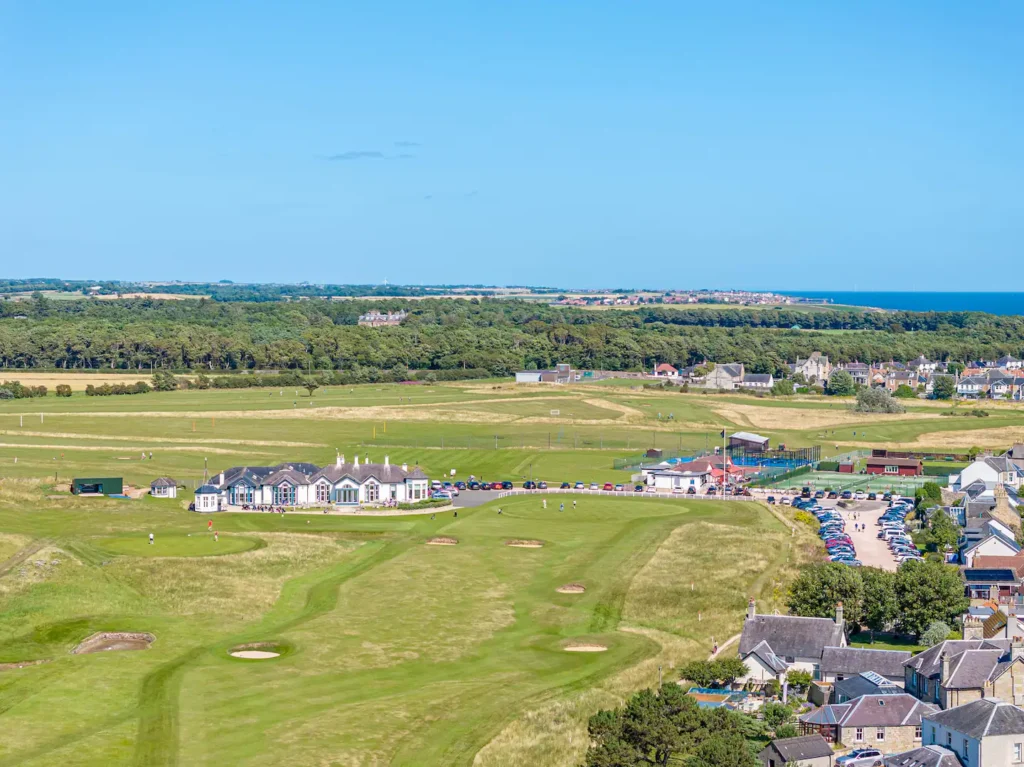 Scenic aerial view of a lush golf course with clubhouse, set against a vibrant blue sky and distant coastal horizon.