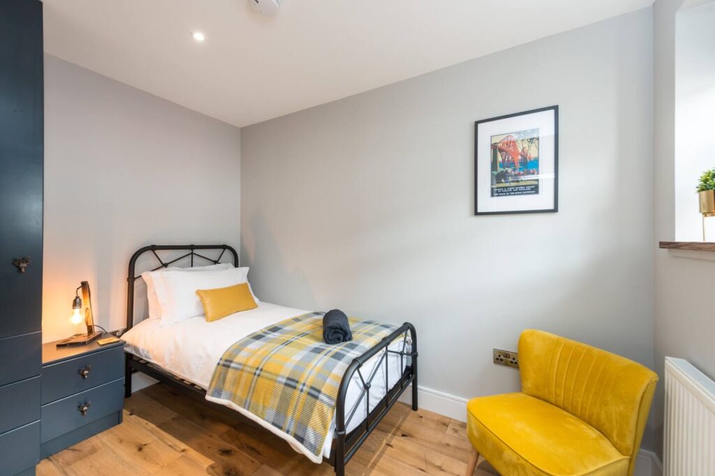 Cozy bedroom with single bed, yellow accents, and a plaid blanket, featuring minimalist decor and a framed wall art.