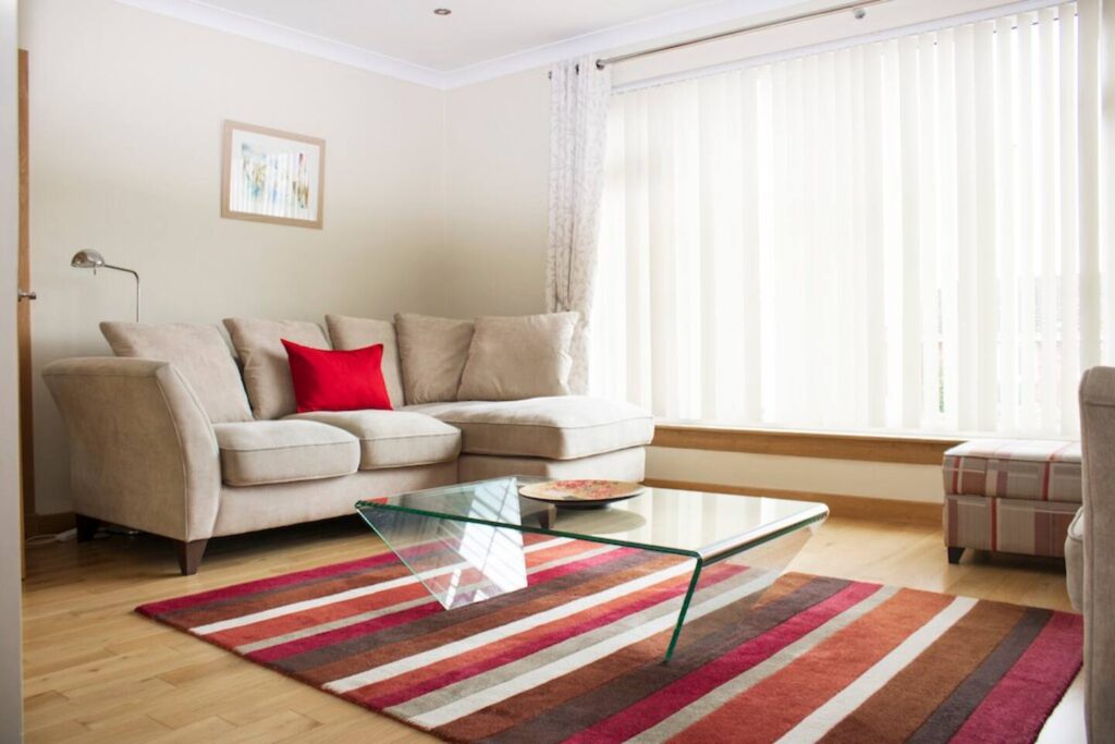 Modern living room with beige sectional sofa, red accents, glass coffee table, and striped rug under soft natural light.