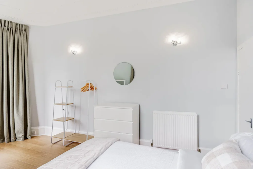 Minimalist bedroom with a light gray theme, featuring a bed, mirror, dresser, and a clothing rack with wooden hangers.