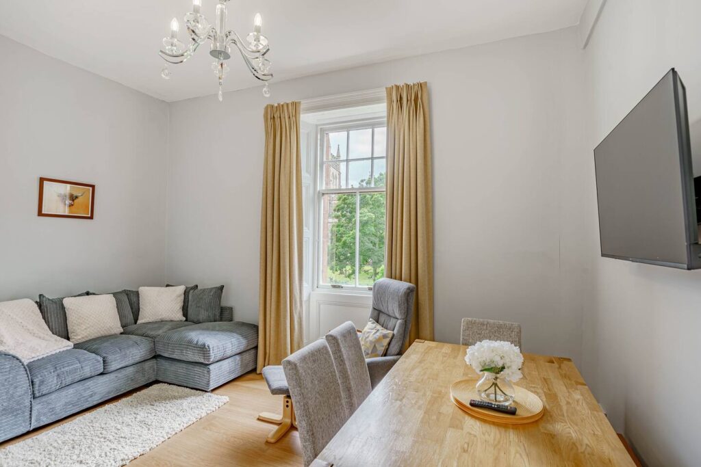 Modern living room with gray couch, dining table, and wall-mounted TV beside a large window with beige curtains.