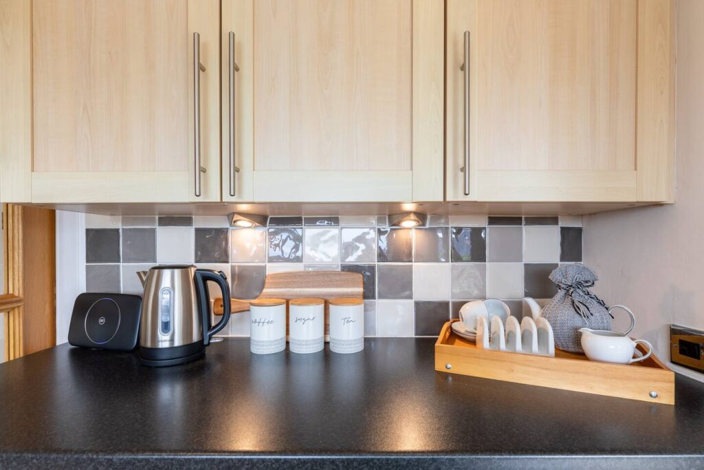 Modern kitchen counter with kettle, storage jars, and dish rack under wooden cabinets and a tiled backsplash.