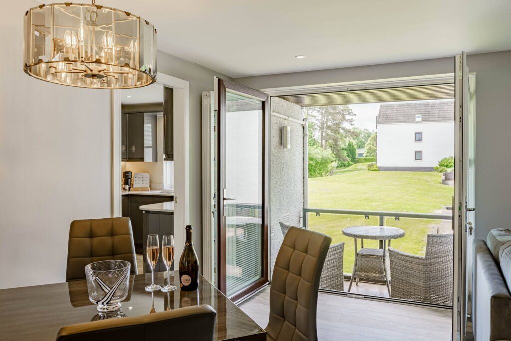 Modern dining area with open doors to a balcony, lush garden view, and a bottle of champagne on the table.