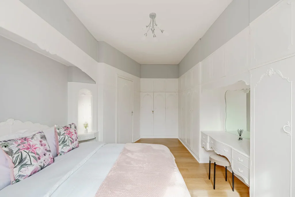 Elegant white bedroom with floral pillows, pink blanket, large closet, and vanity table on wooden floor.